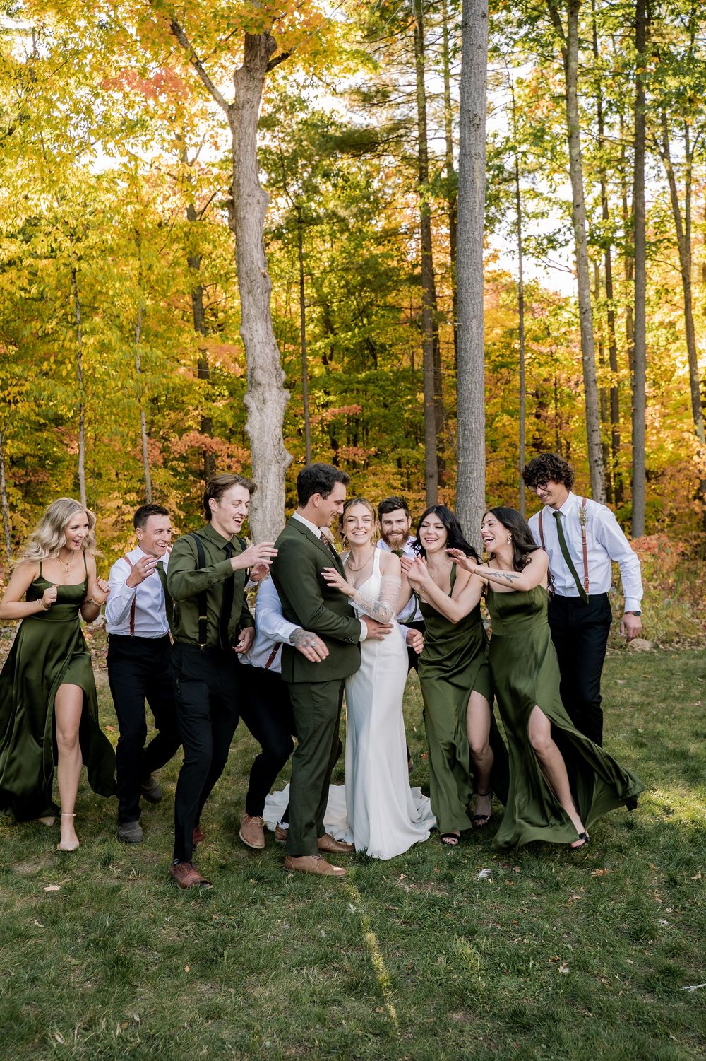 Wedding party poses playfully in a line against autumn trees with golden foliage in the background.