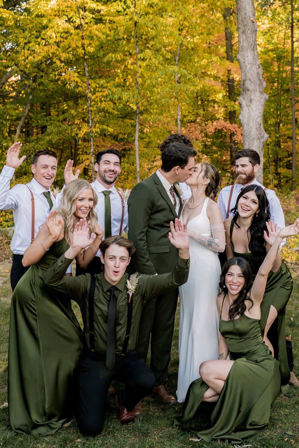Wedding party in olive green and white attire poses playfully in a forest setting with golden autumn foliage.