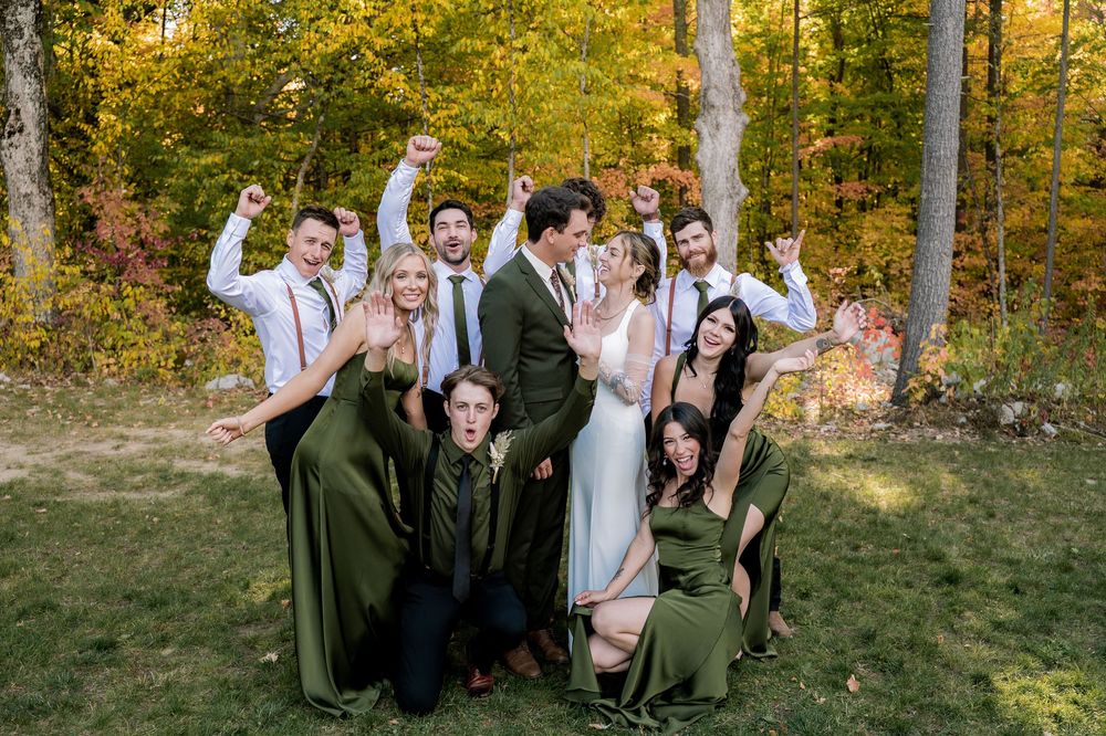 Wedding party in green and white attire poses playfully together on grass with autumn foliage in background.