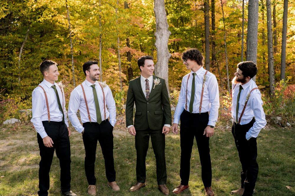 A group of groomsmen in suspenders and ties stand together in a sunlit autumn forest with golden foliage.