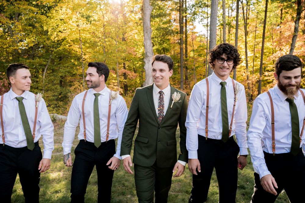 Groomsmen in white shirts and suspenders stand together against a backdrop of golden autumn trees in sunlight.