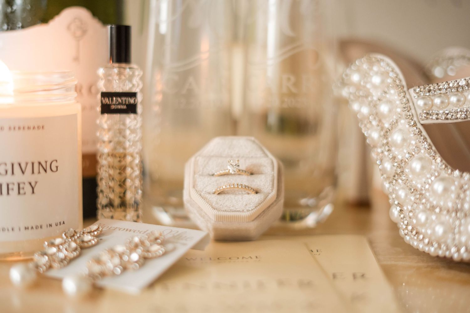 Elegant wedding flat lay with diamond rings in velvet box, pearl jewelry, Valentino perfume, and candle on gold surface.