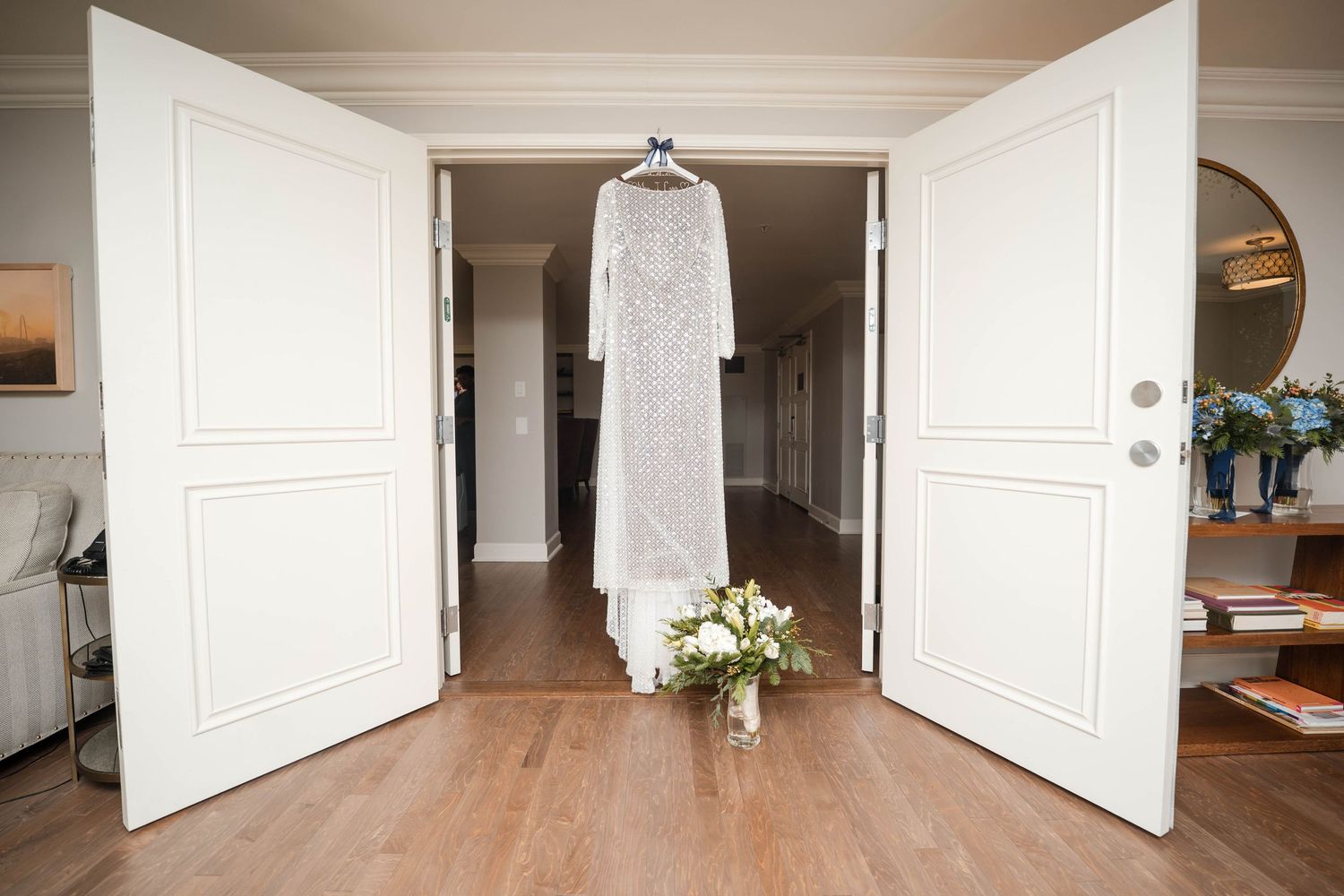 White lace wedding dress hanging between open double doors with a small bouquet below.