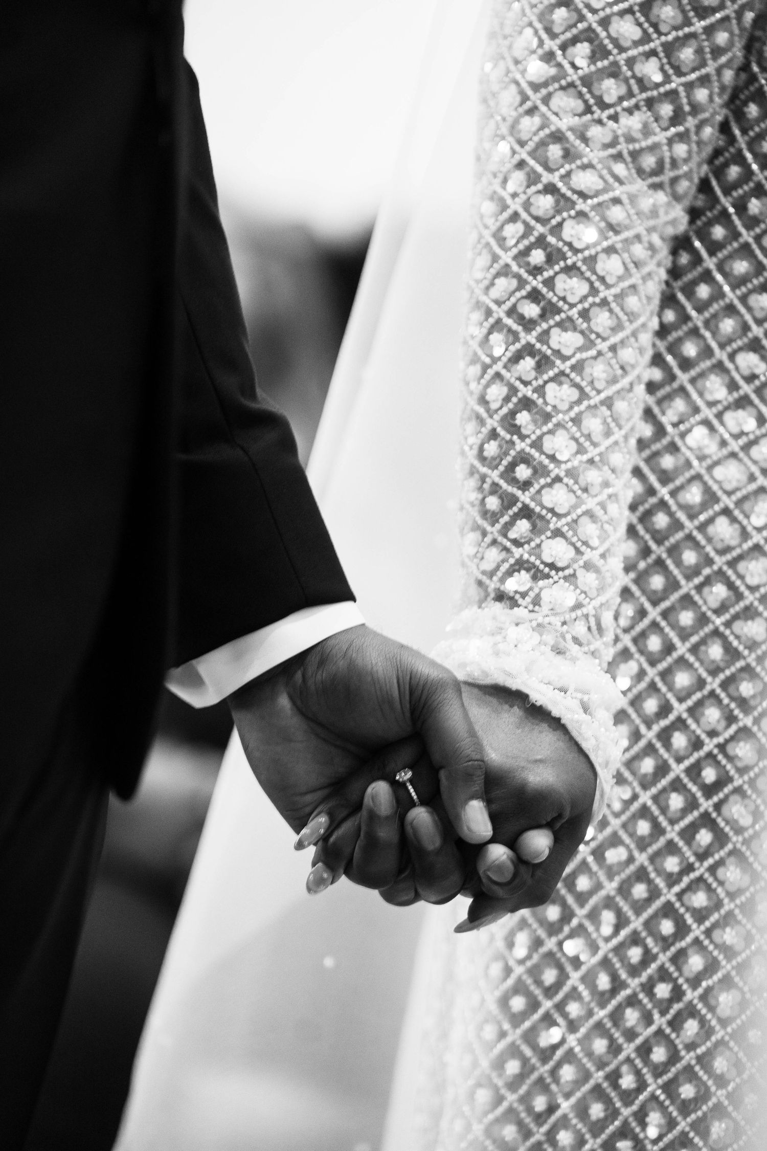 Close-up black and white photo of newlyweds holding hands showing diamond engagement ring and lace wedding dress.