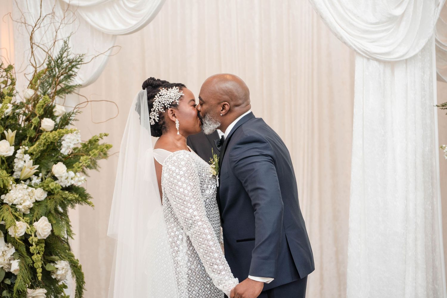 Bride and groom sharing first kiss at altar decorated with white floral arrangements and draped fabric backdrop.