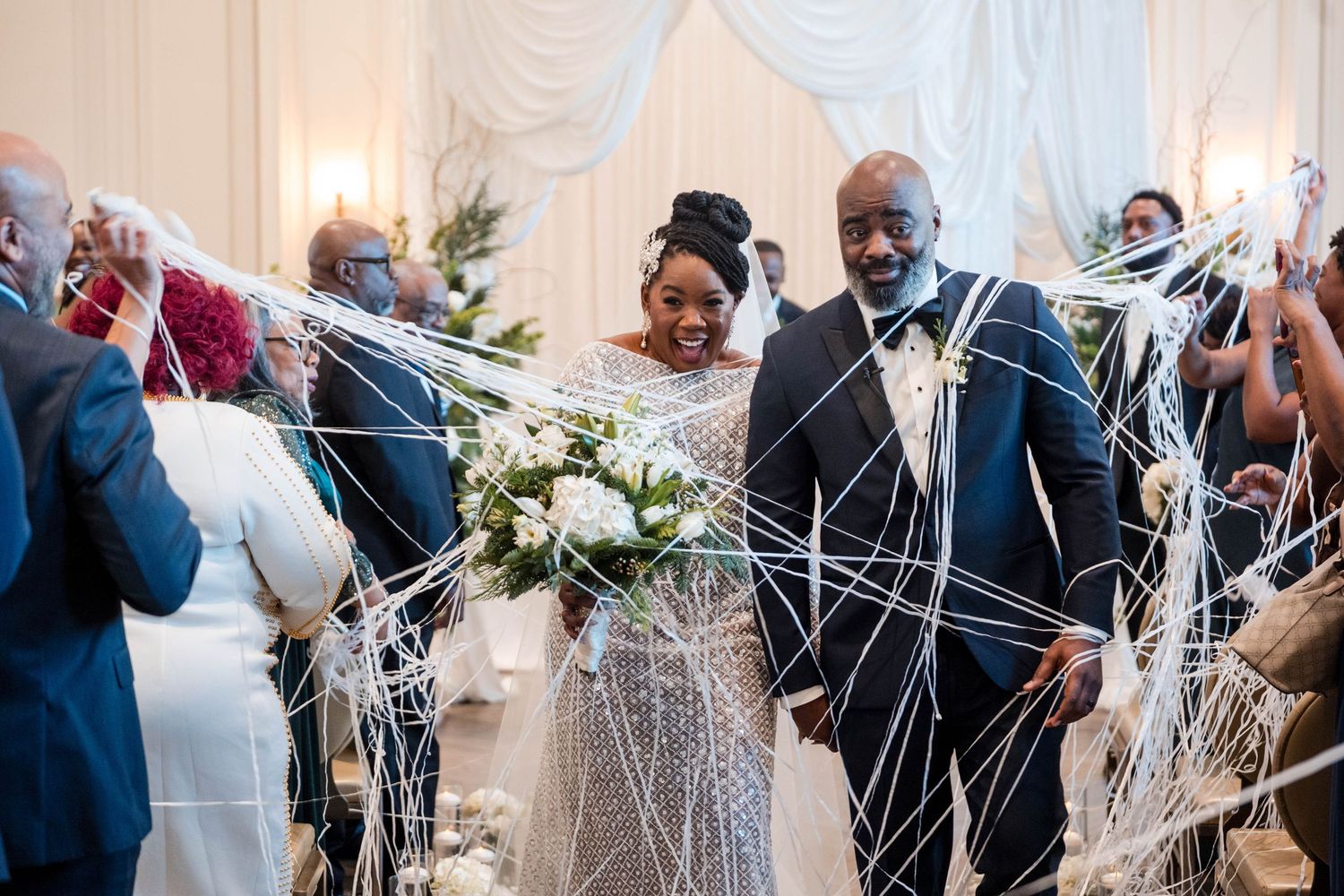 Joyful newlyweds walking down aisle surrounded by guests throwing white ribbon streamers at elegant wedding ceremony.