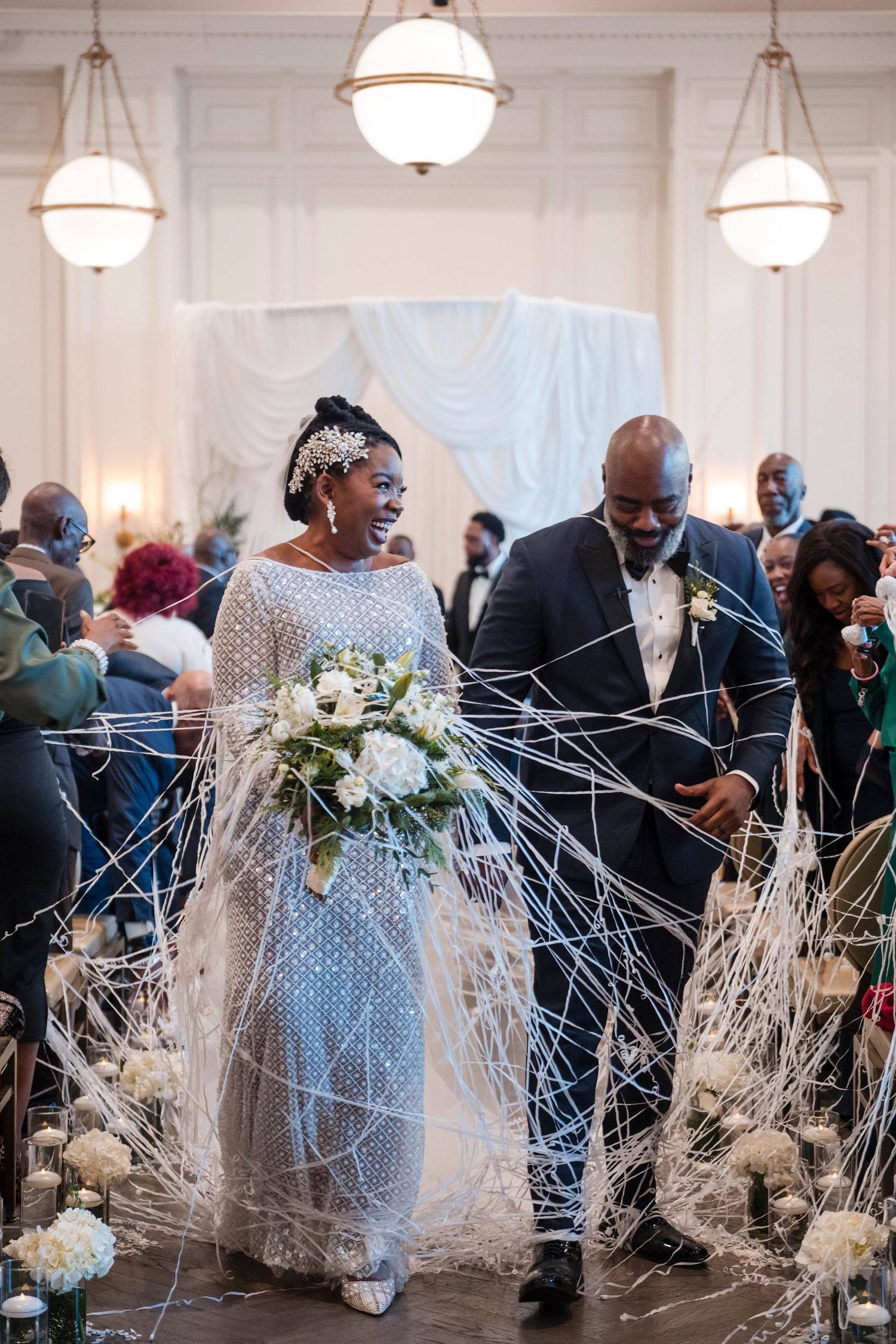 Smiling bride in beaded gown and groom exit ceremony aisle through white ribbon streamers thrown by guests.