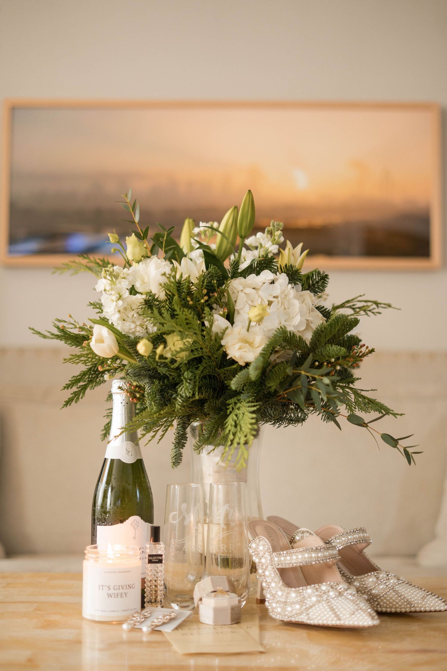 Elegant wedding flat lay with white floral bouquet, champagne bottle, heels, and candle.