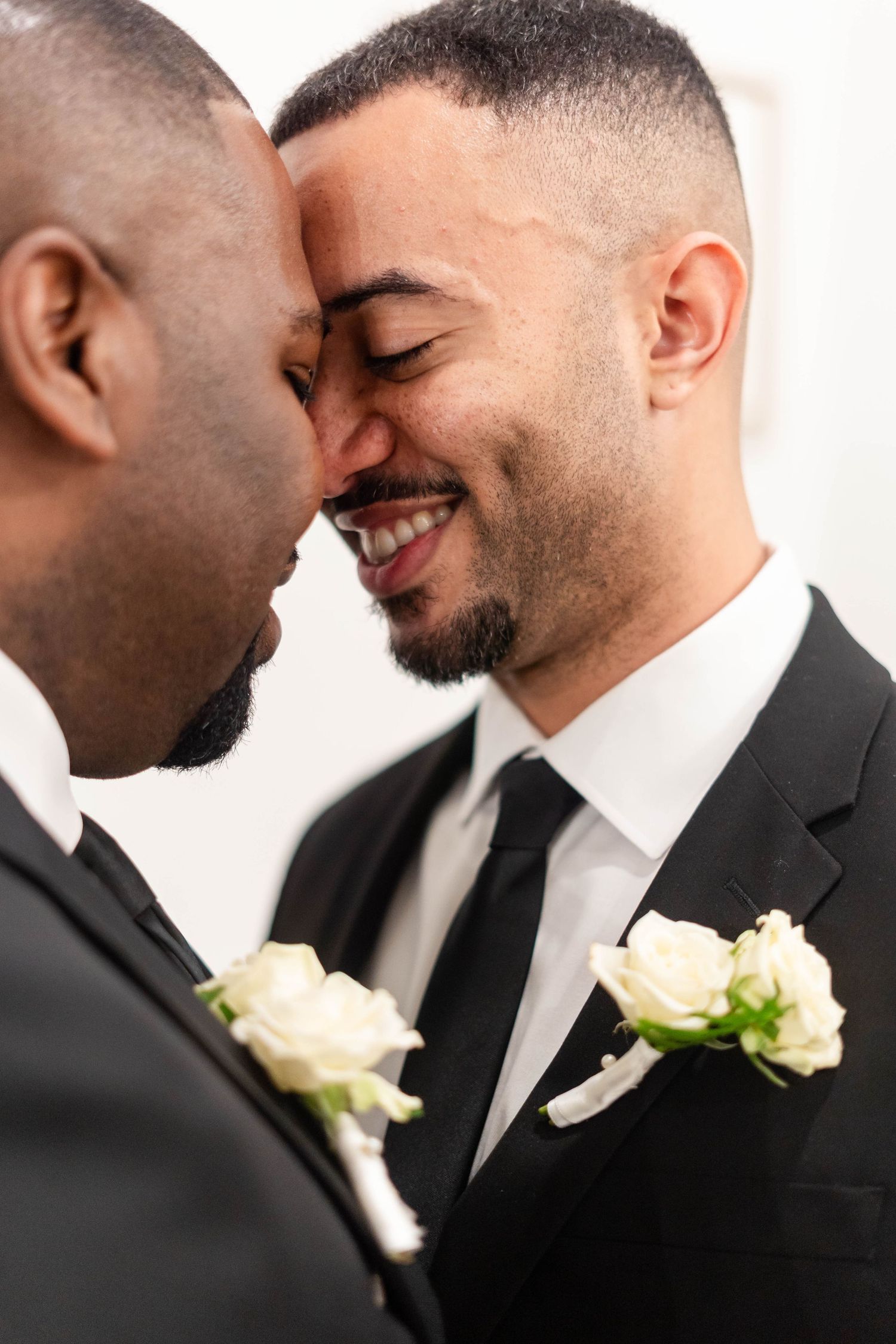 Two grooms in black suits with white rose boutonnieres share an intimate forehead touch moment on their wedding day.