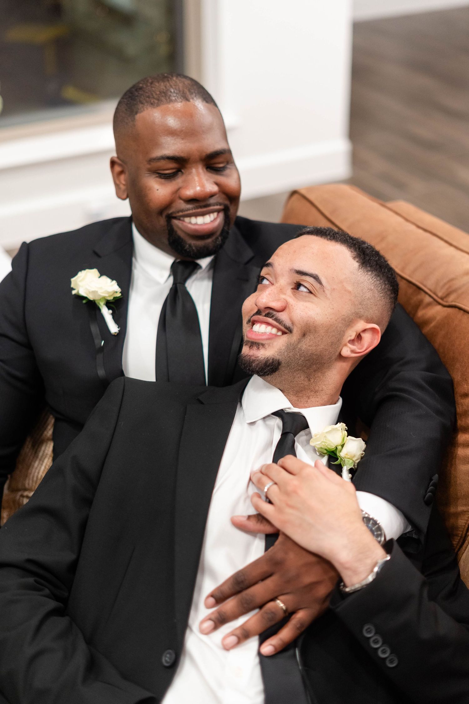 Two grooms in matching black suits with white boutonnieres share a joyful embrace while seated together.