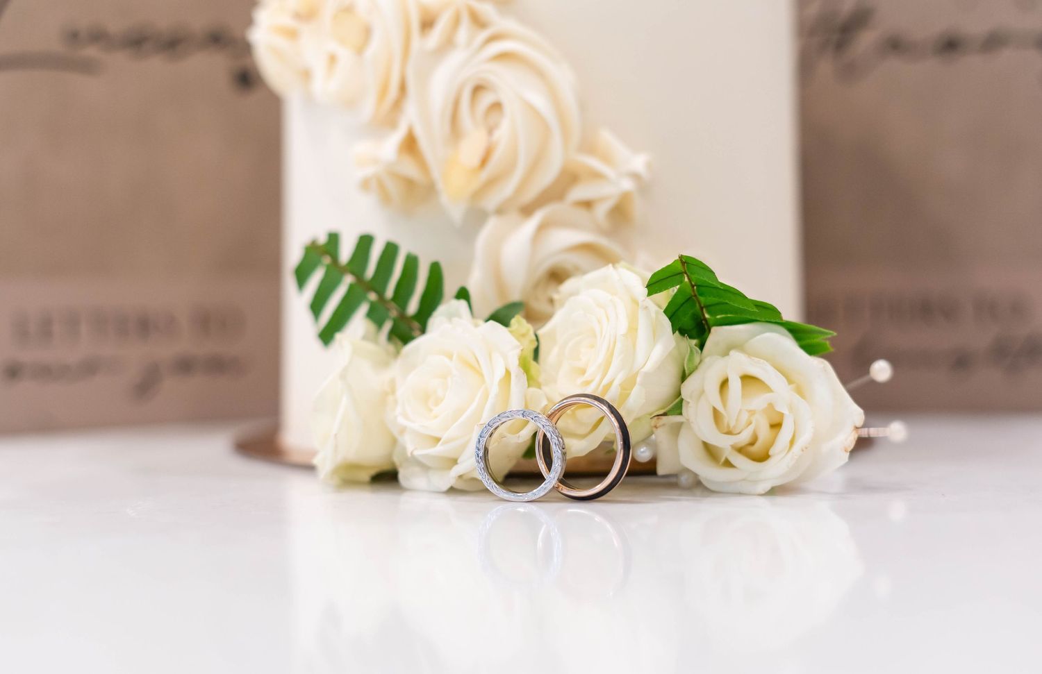 Two silver wedding bands resting against white roses beside a rosette wedding cake on white surface.
