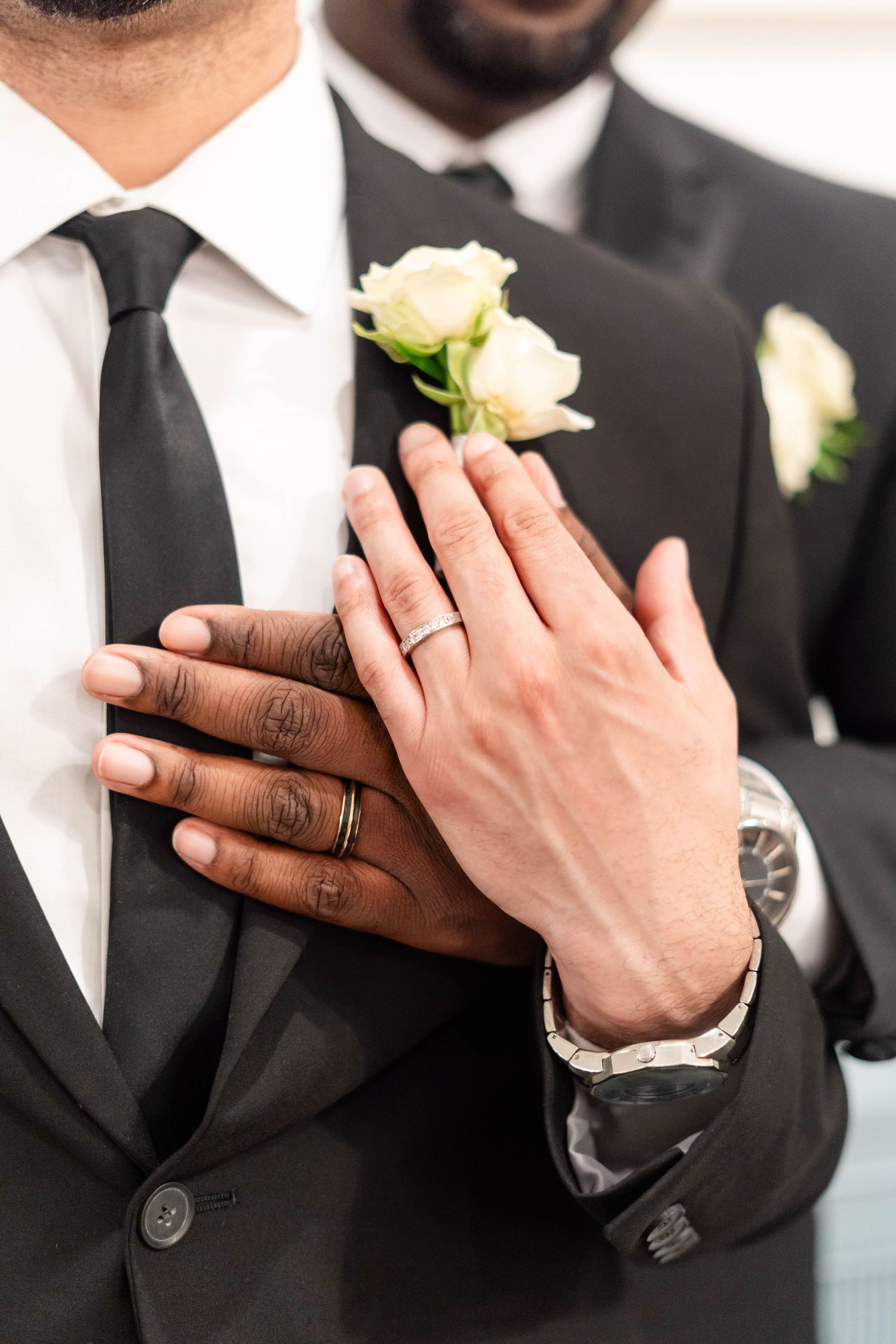 Close-up of interracial groom couple holding hands showing wedding bands with white rose boutonnieres on black suits.