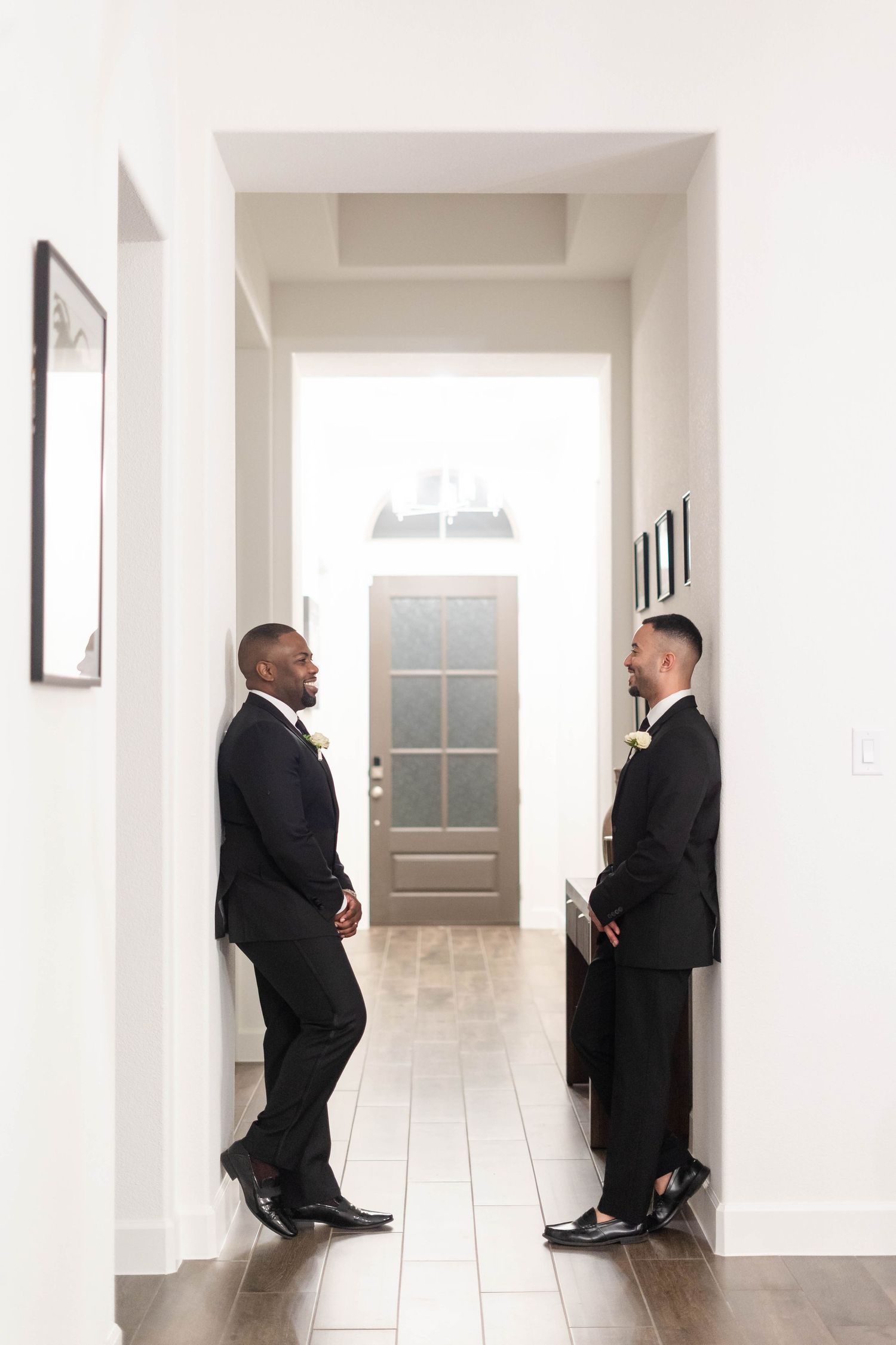 Two grooms in matching black suits with white rose boutonnieres standing in bright white hallway before wedding.