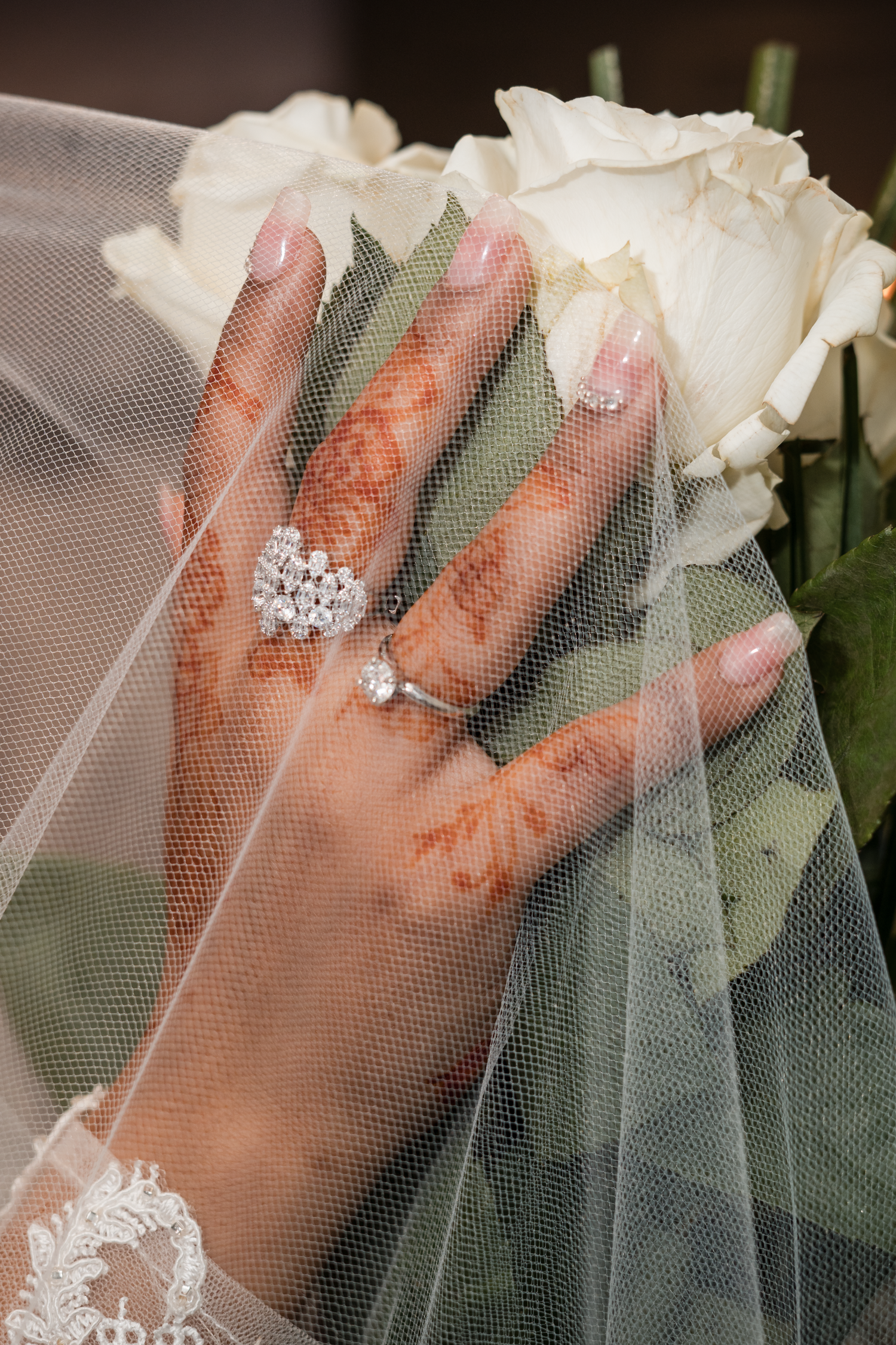 Bride's hand with henna, diamond engagement ring, and pearl ring resting on white roses.