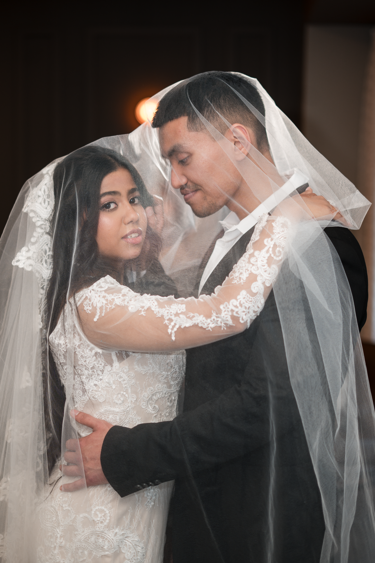 Bride and groom embracing tenderly under white lace veil in warmly lit wedding portrait.