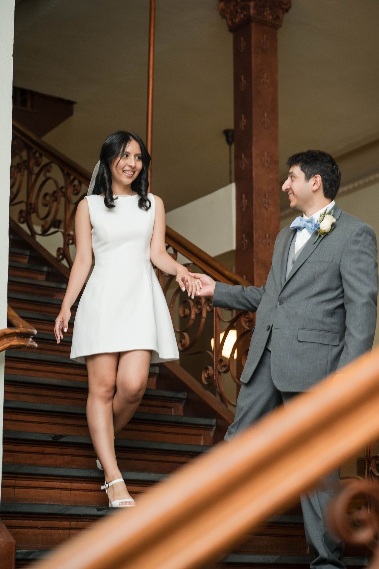 Bride in white mini dress and groom in gray suit hold hands on ornate wooden staircase.