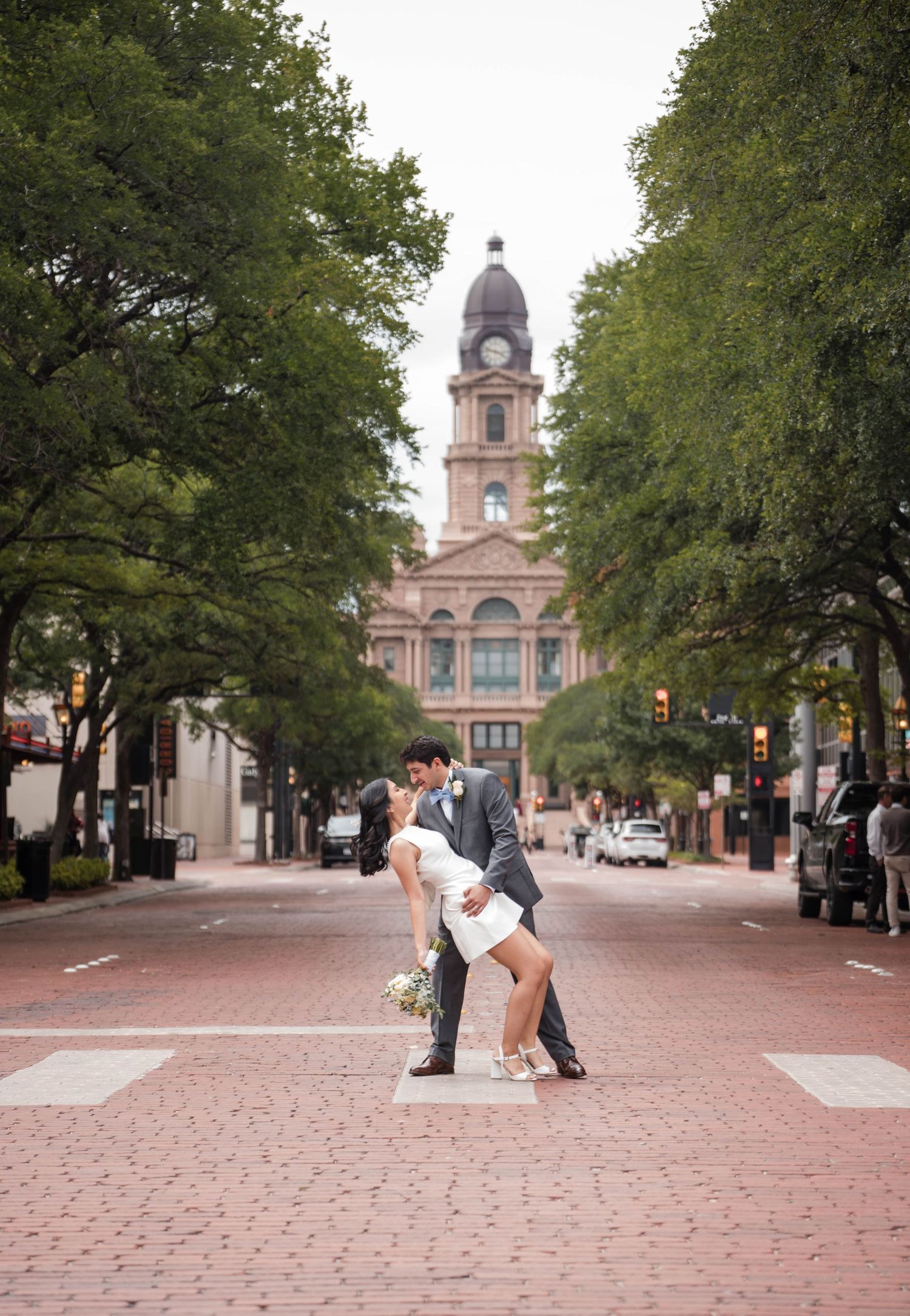 Couple shares romantic dip kiss on brick street with historic courthouse backdrop surrounded by lush trees.