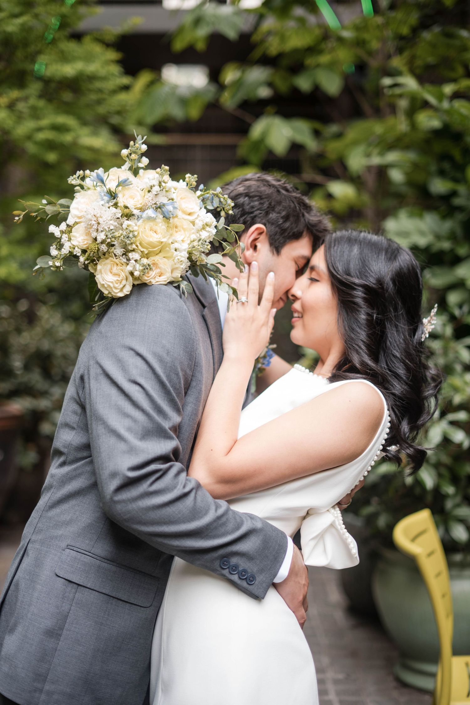 A bride and groom share a romantic moment, nearly kissing, as she holds a white and blue floral bouquet outdoors.