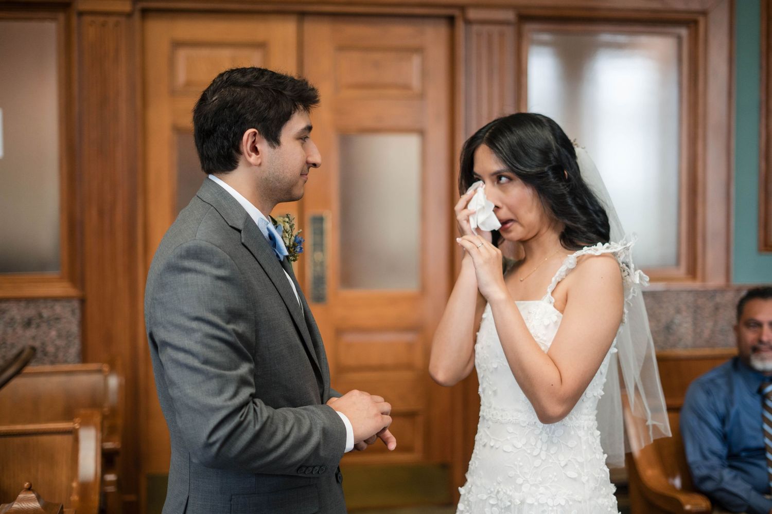Emotional bride wiping tears during wedding ceremony while groom holds her hand in elegant venue.