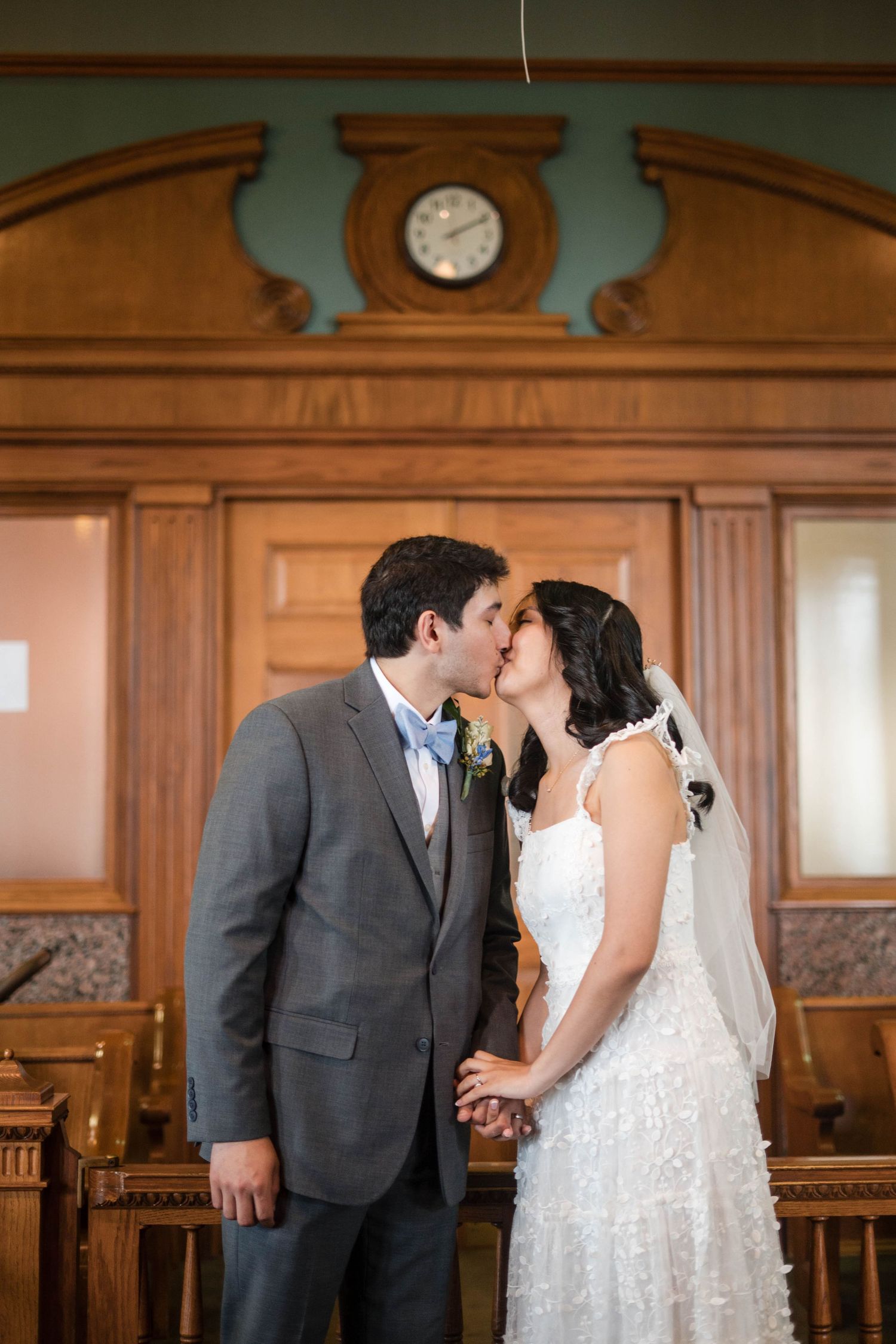 Bride and groom share a kiss at their wedding ceremony in an elegant wood-paneled venue with a clock backdrop.