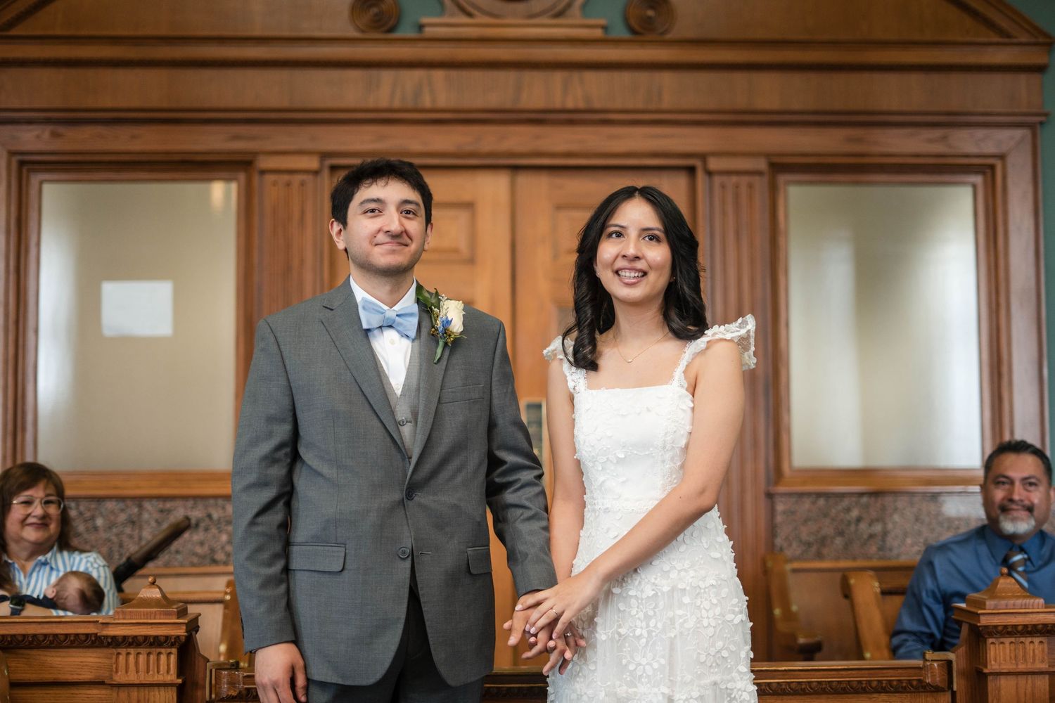 Smiling newlywed couple holding hands at courthouse wedding ceremony, groom in gray suit, bride in white lace dress.