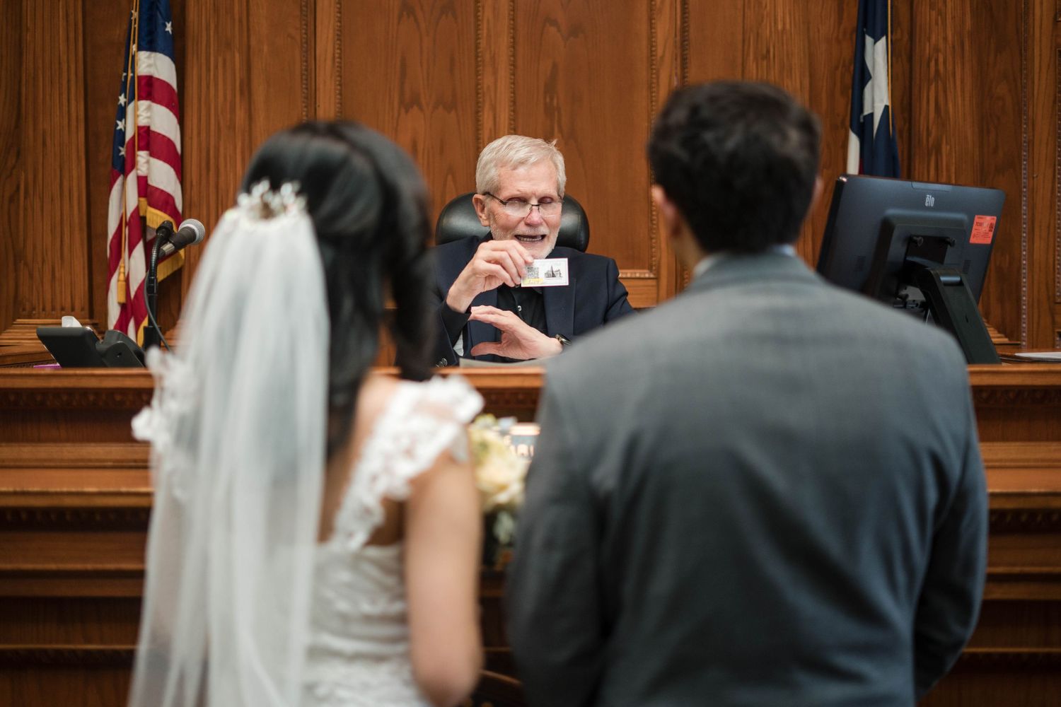 A judge officiates a wedding ceremony for a couple in bridal attire inside a courtroom with American flags.