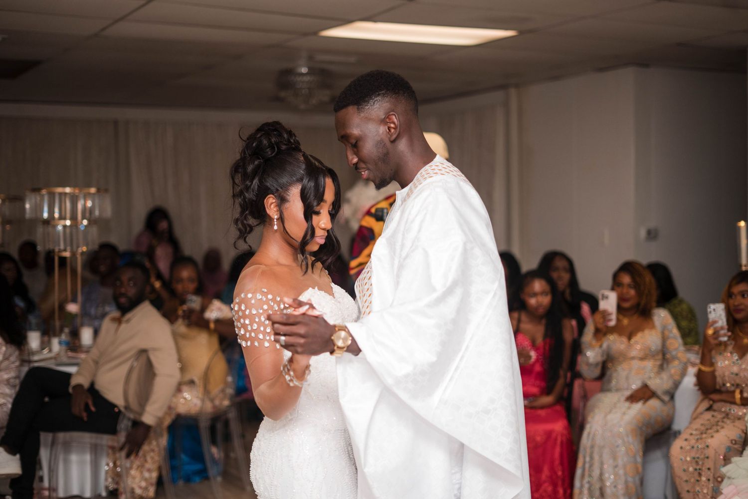 Bride and groom share first dance as guests watch at wedding reception, groom in white African attire.