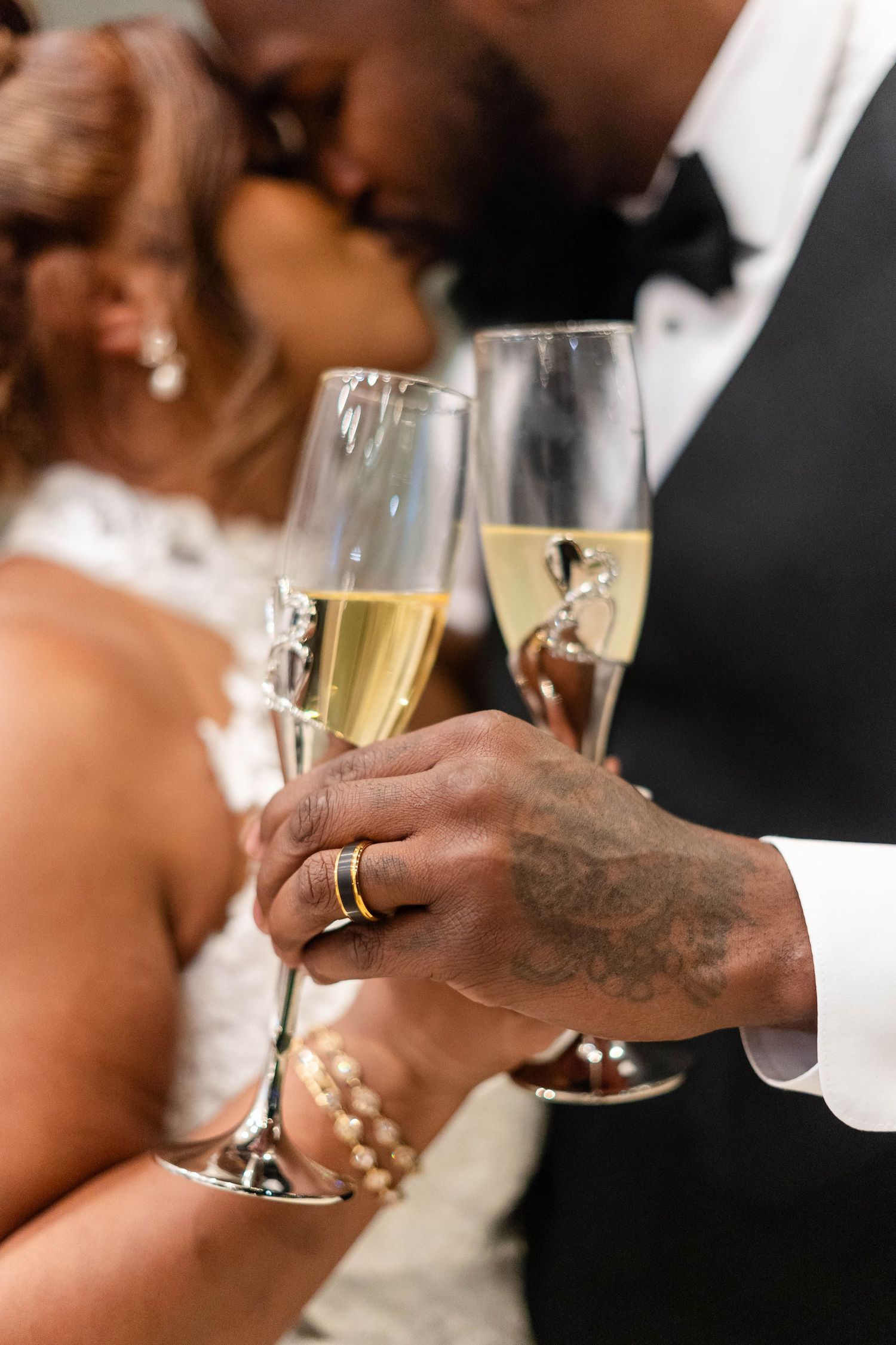 Close-up of tattooed groom and bride clinking champagne glasses with wedding rings visible.