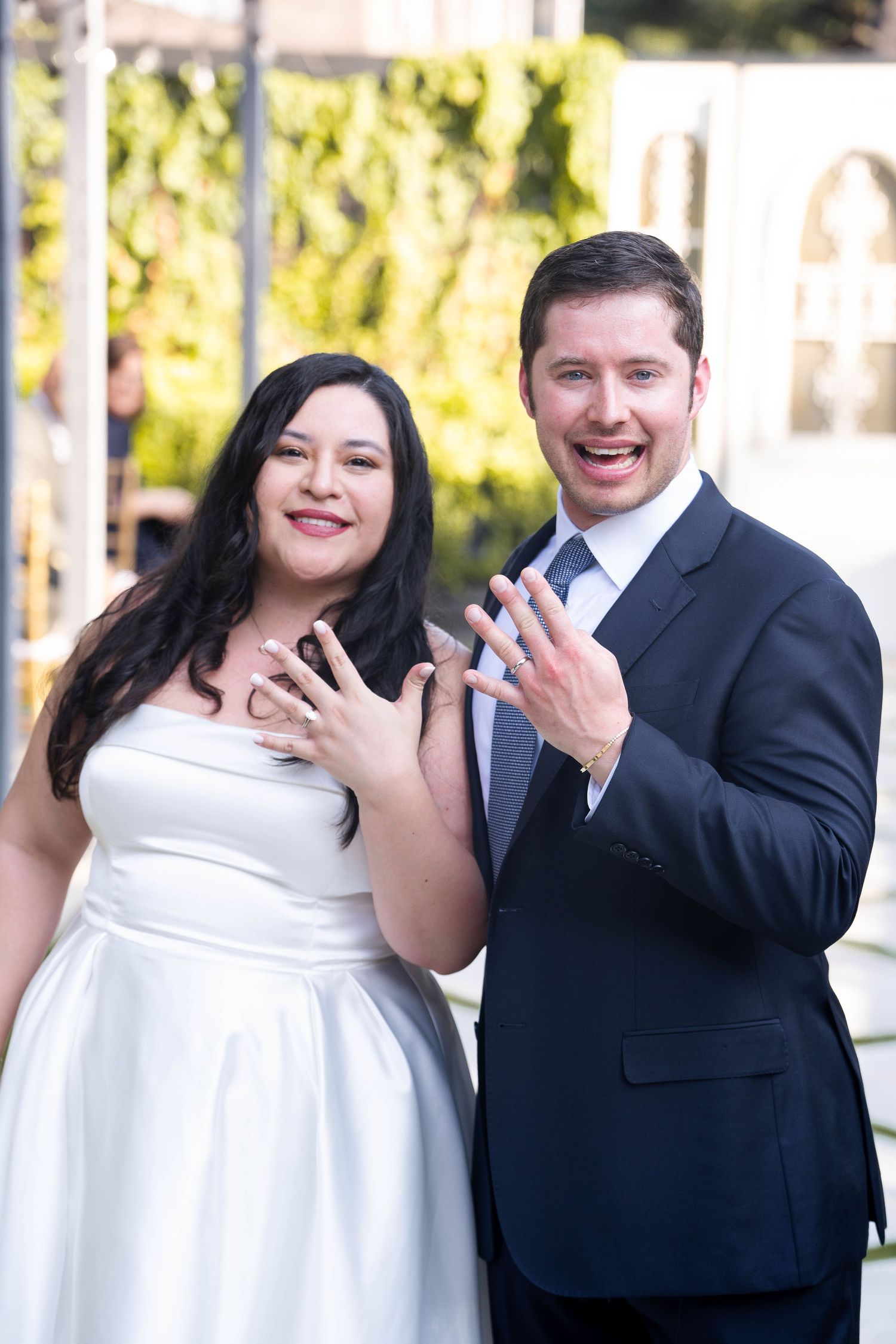 Smiling newlywed couple showing off wedding rings outdoors after ceremony.