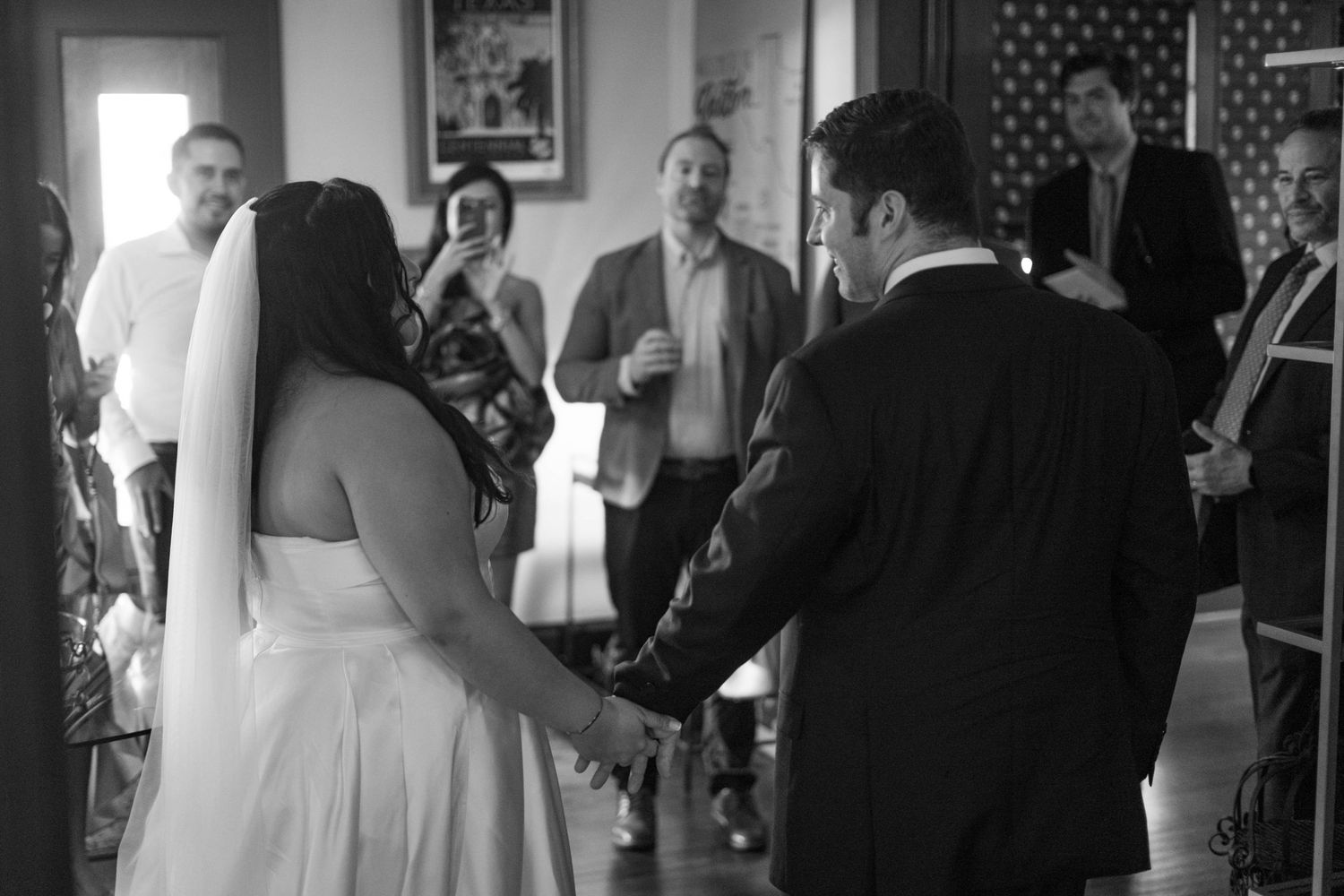 Black and white photo of bride and groom holding hands during indoor wedding ceremony with guests watching.