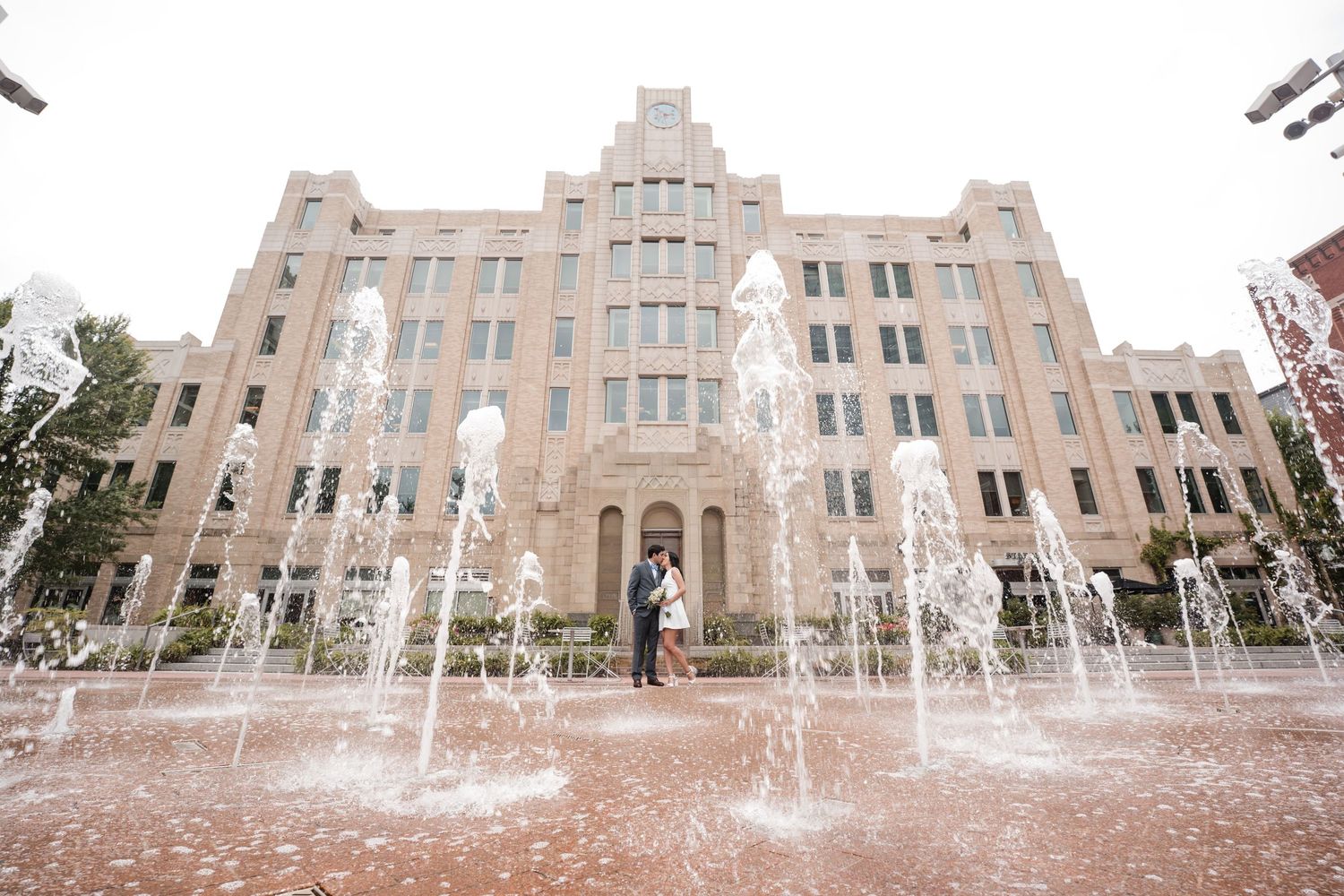 Couple embracing among fountain jets in front of a grand beige stone building on a brick plaza.