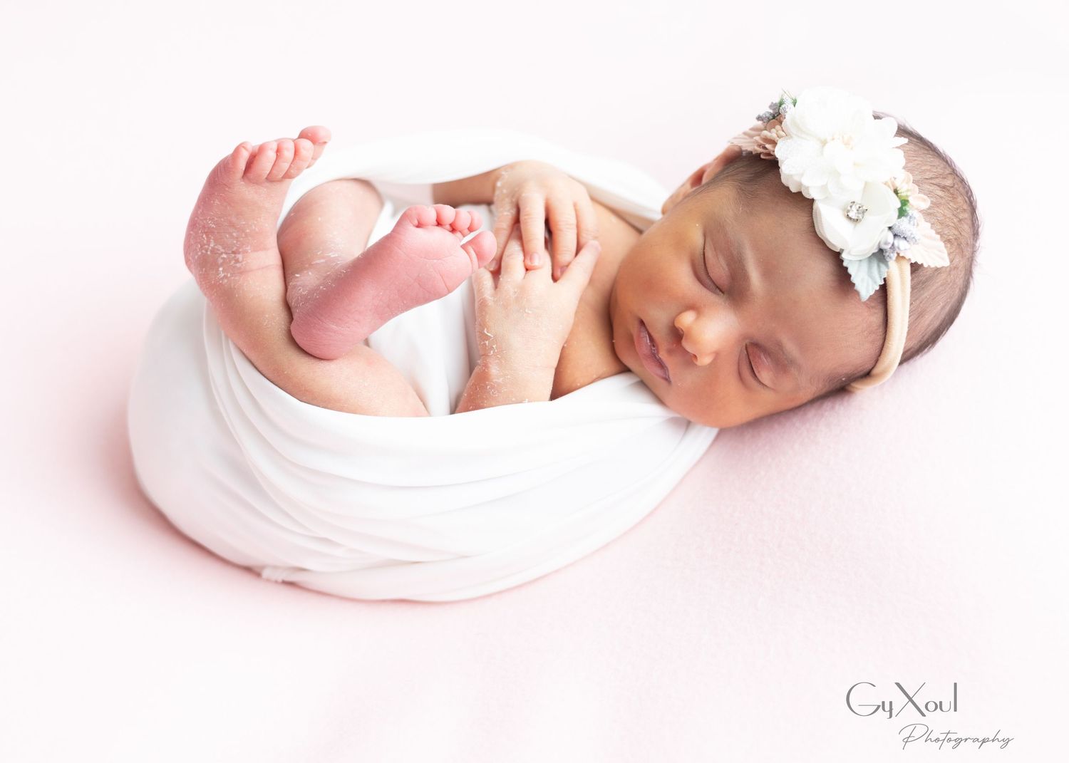 A newborn baby wearing a white headband lies on a pink background in a delicate pose during a professional photo session.