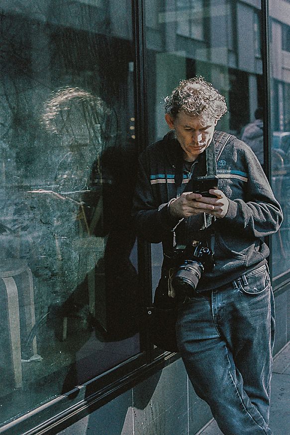 Person in denim outfit leaning against window while looking at phone on urban street.