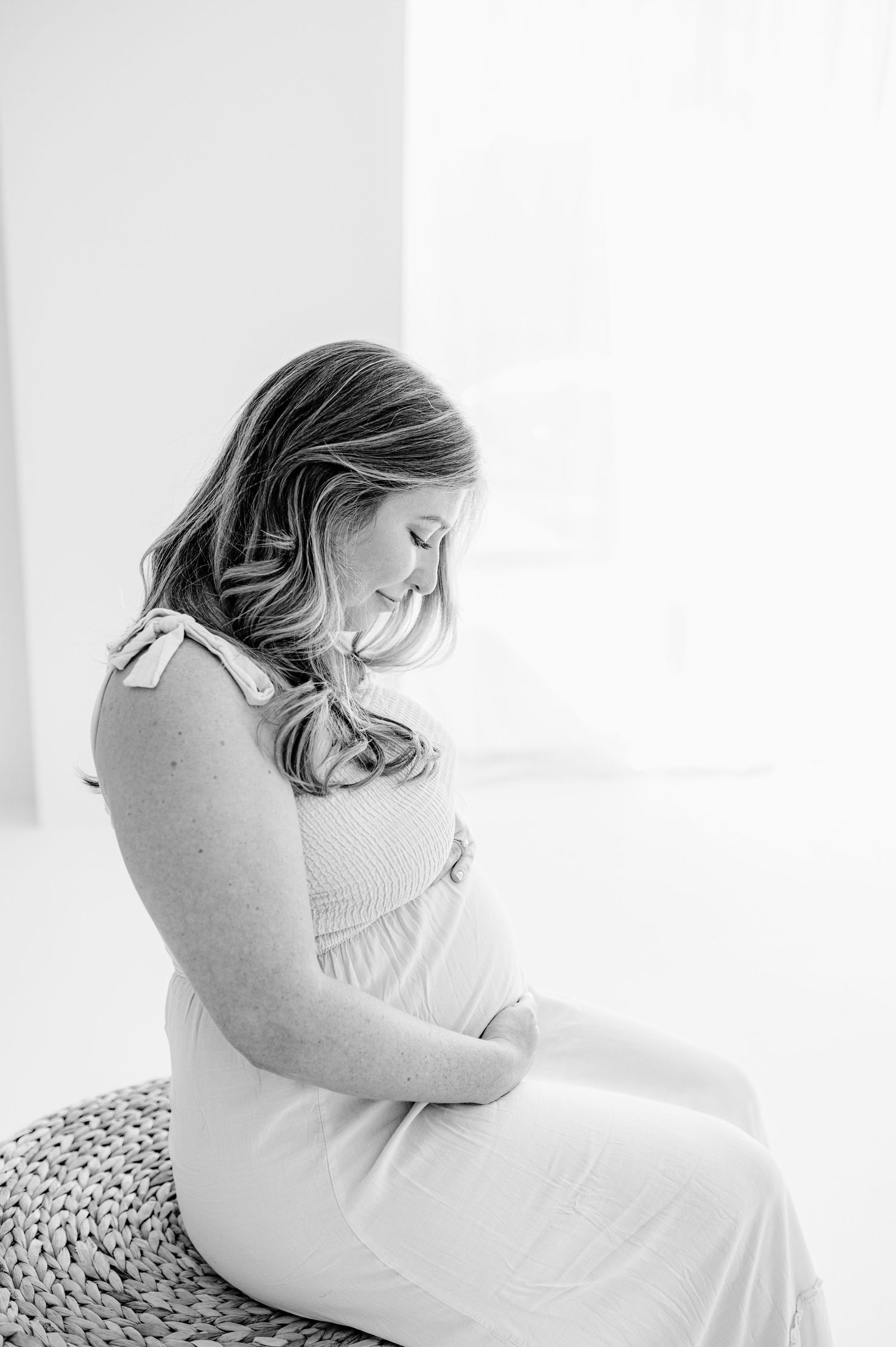 Pregnancy photography in Jacksonville, FL showing a pregnant woman in white dress sitting in soft window light.
