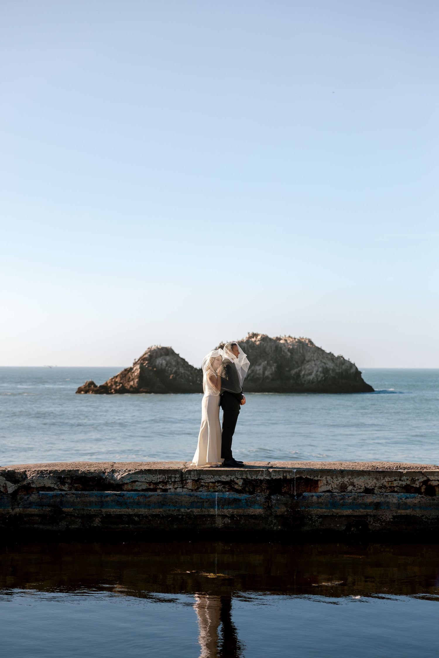 Silhouetted couple stands on coastal seawall in full sun with rocky islands visible in the ocean background at Sutro Baths in San Francisco