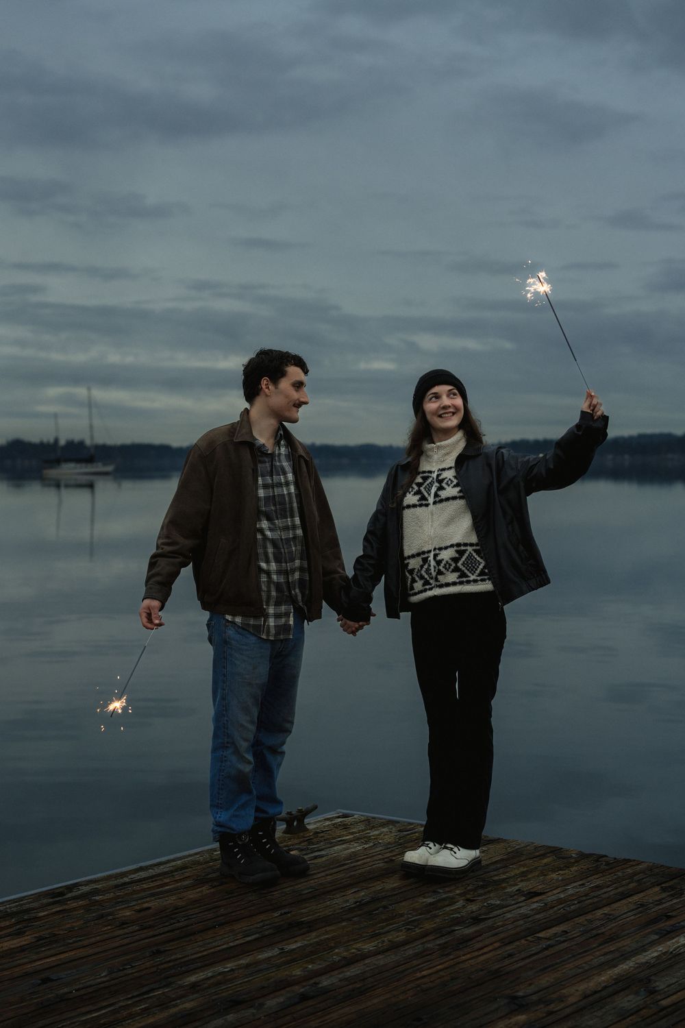 Two people hold sparklers while standing on a wooden dock at dusk overlooking a misty waterfront.