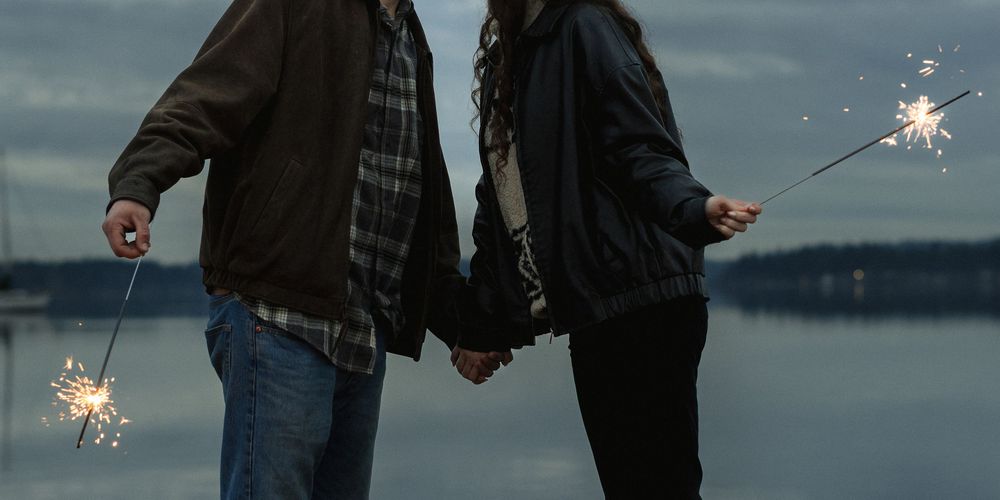 A couple holds hands while holding sparklers at dusk by a serene lake.