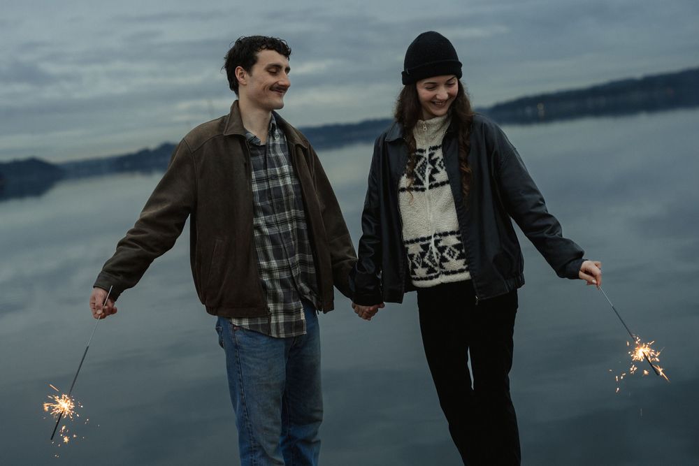 Two people hold hands and sparklers while standing near a lake at dusk in a moody outdoor setting.