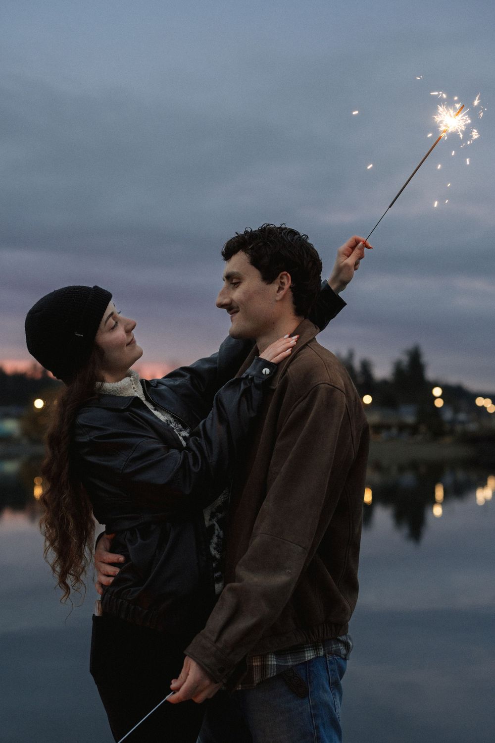 A romantic couple holds sparklers by a reflective lake at dusk with city lights twinkling in the background.
