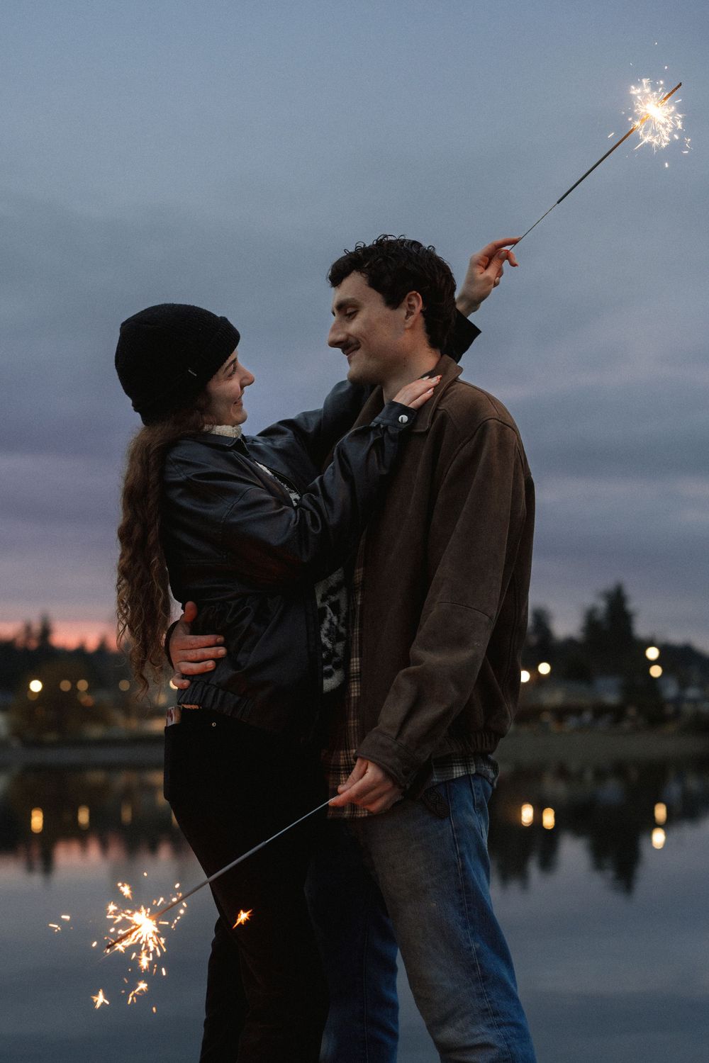 A couple shares an intimate moment while holding sparklers near a lake during twilight hours.