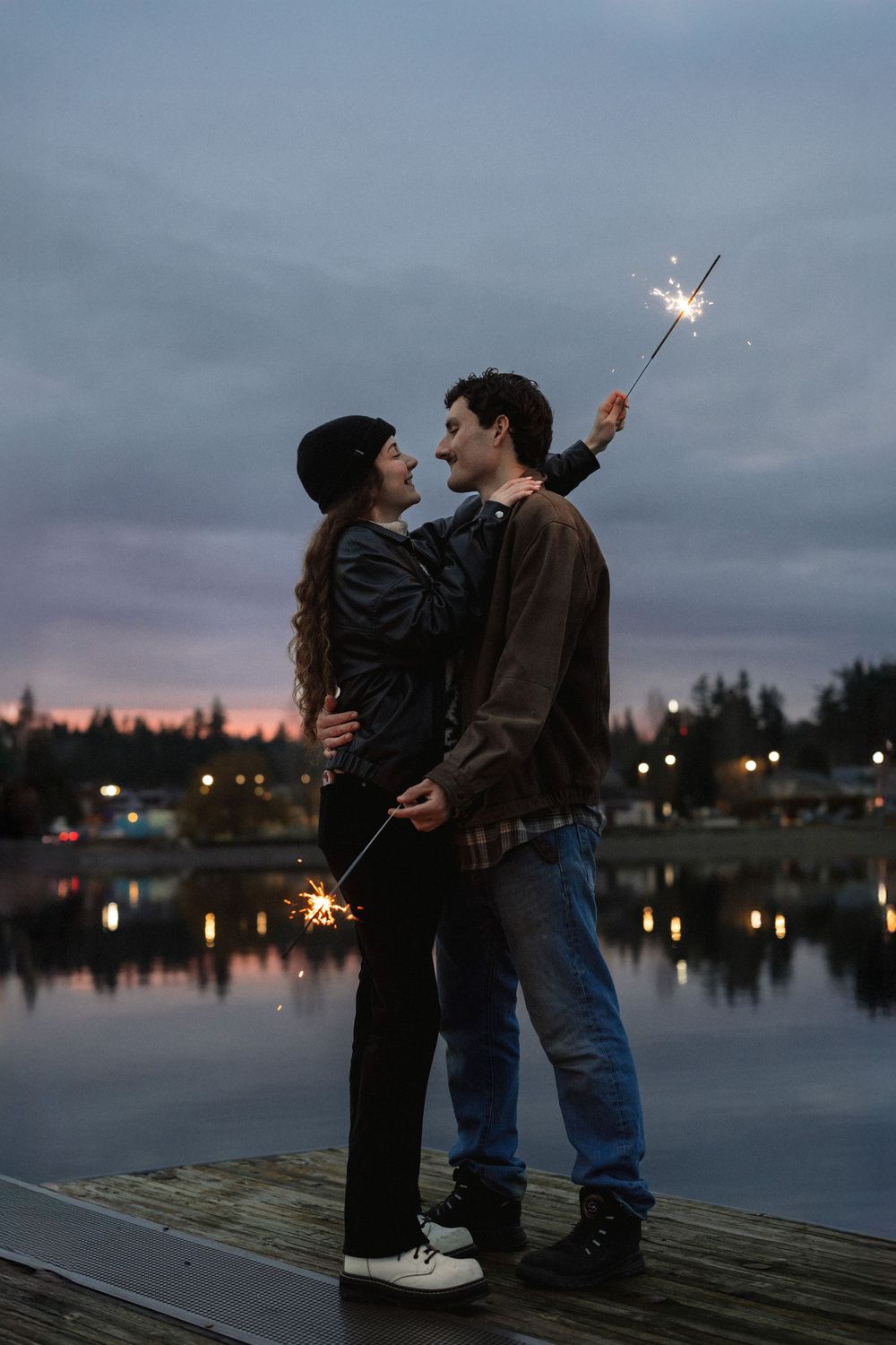 A couple shares a romantic moment on a dock at twilight while holding sparklers over a reflective lake.