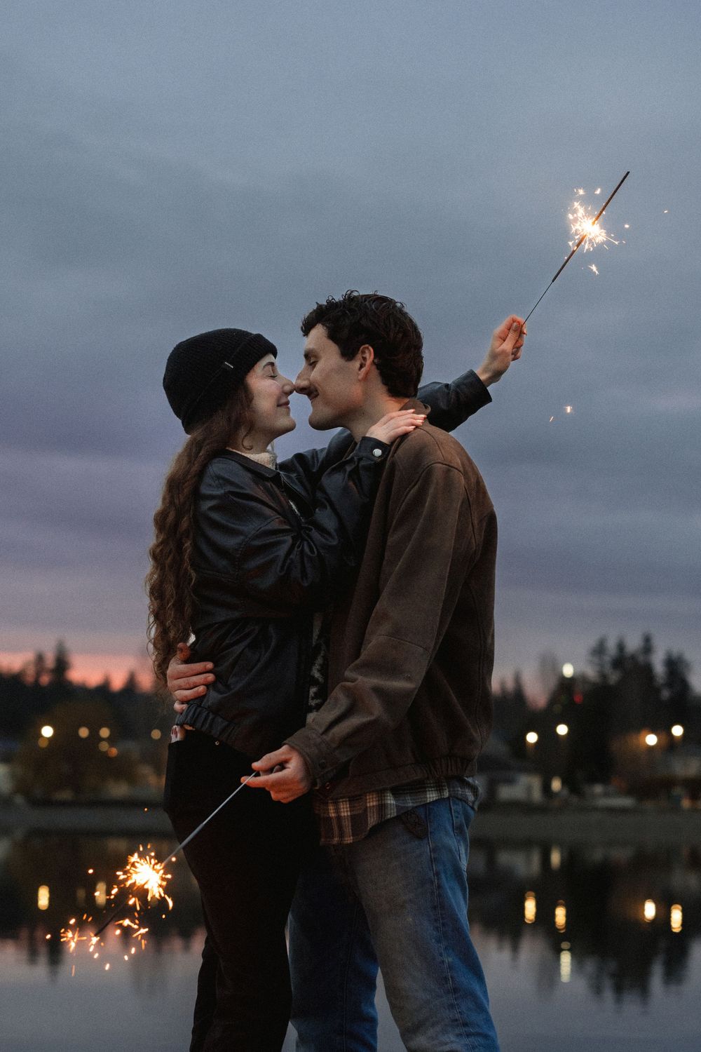 A romantic couple holds sparklers while embracing by a lake at dusk with city lights reflecting on the water.