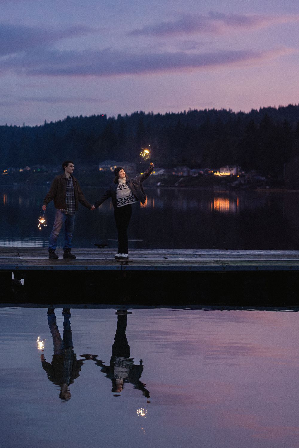 People holding sparklers create reflections on a tranquil lake at dusk with twinkling lights in the background.