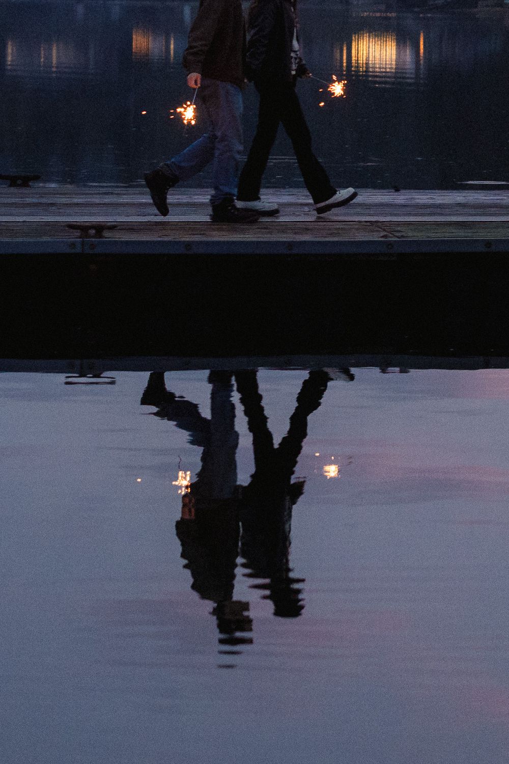 Dark silhouettes cast reflections on a wet surface during twilight with distant lights creating an atmospheric scene.