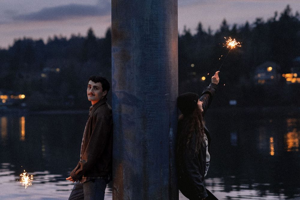 Two people celebrate with sparklers against a twilight waterfront backdrop with city lights in the distance.