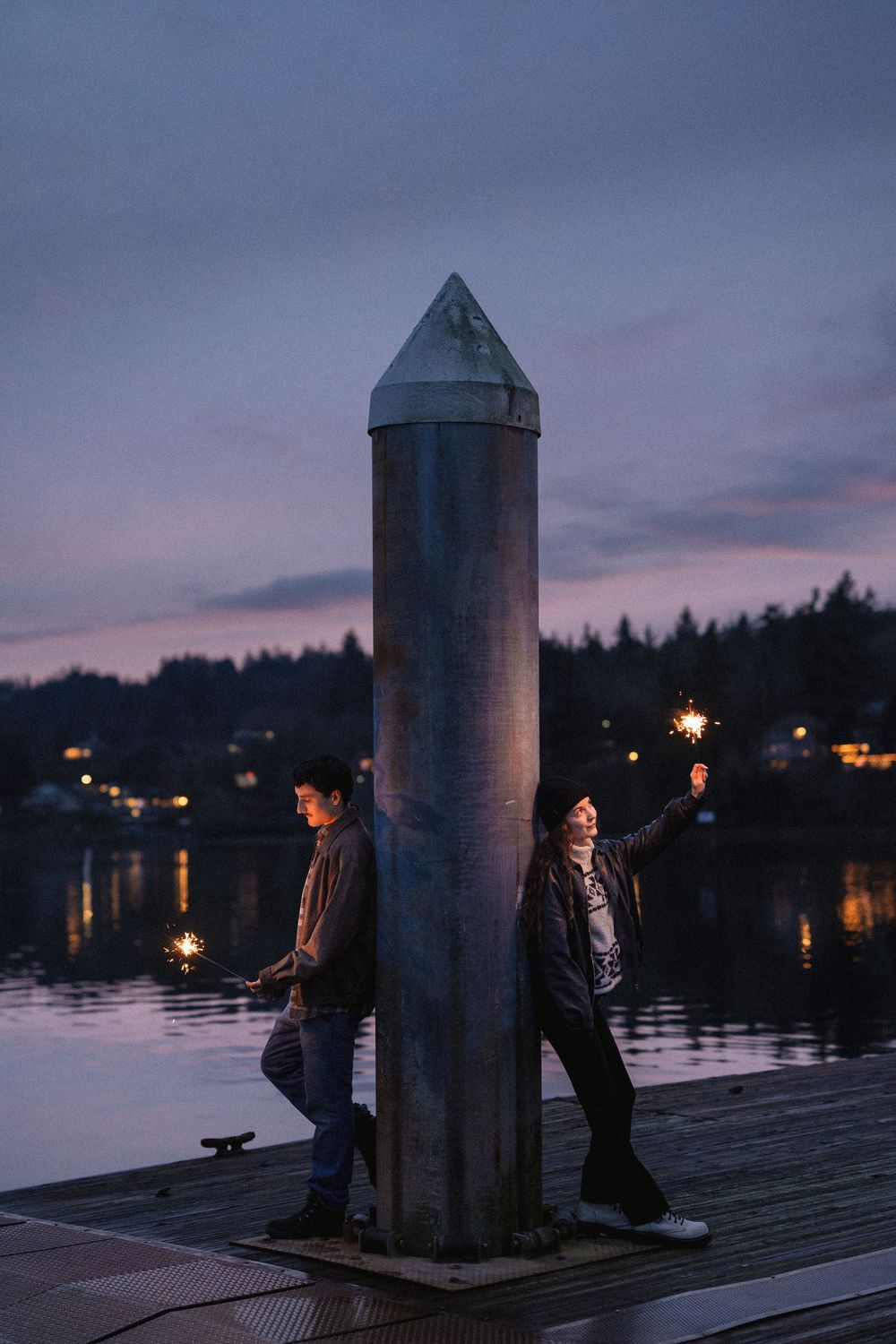 Two people lean against a tall pillar on a waterfront dock at dusk with city lights twinkling in the background.