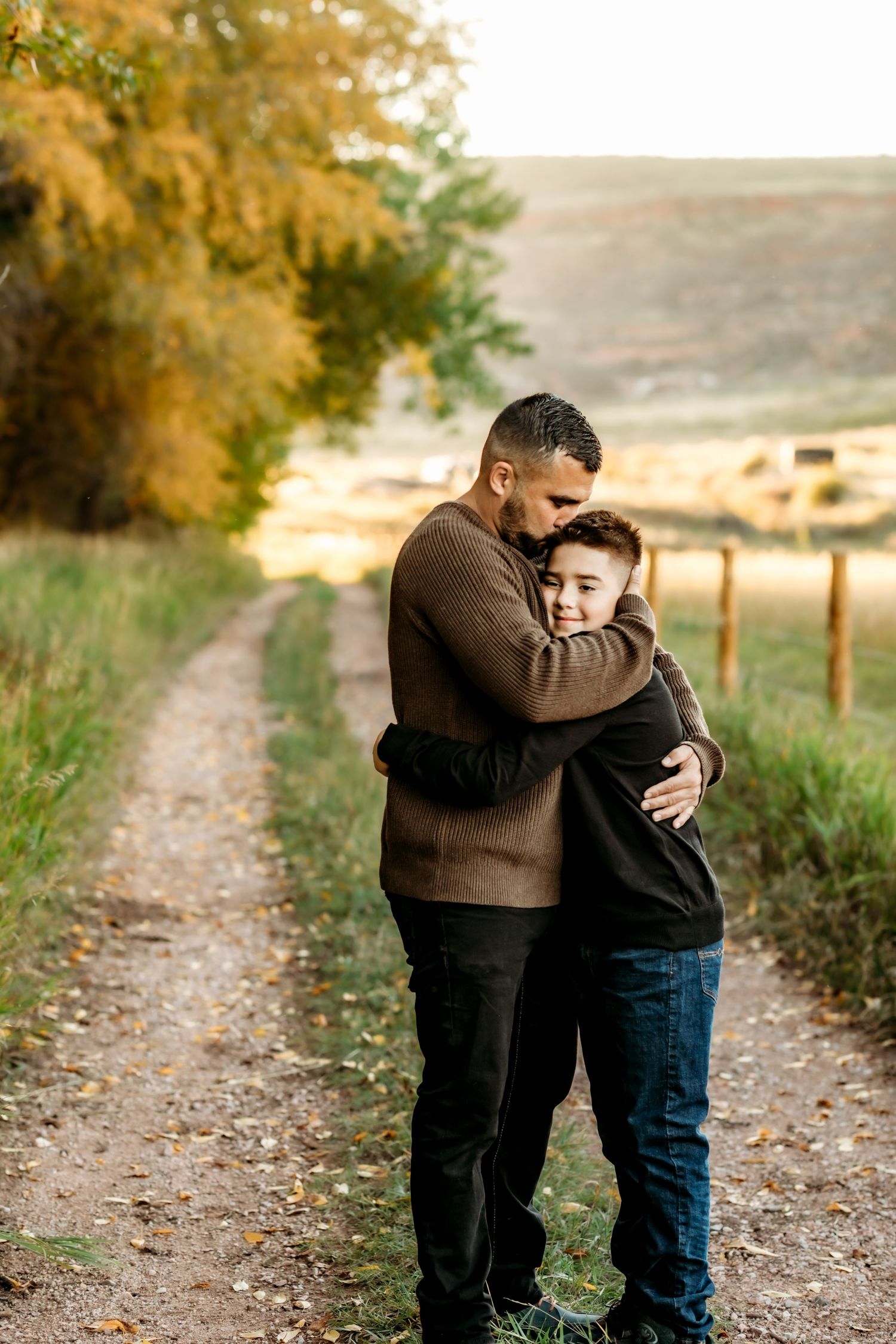 Two people embrace tenderly on a dirt path surrounded by autumn foliage and golden countryside scenery.