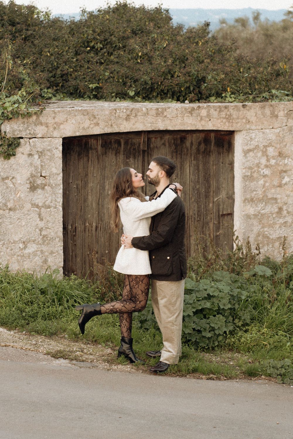 A romantic couple embracing against a rustic stone wall and wooden gate in a natural setting.