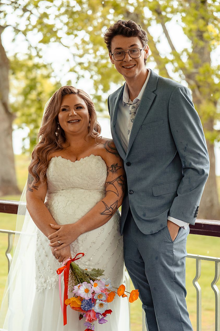 A couple in wedding attire poses together outdoors with autumn foliage in the background.