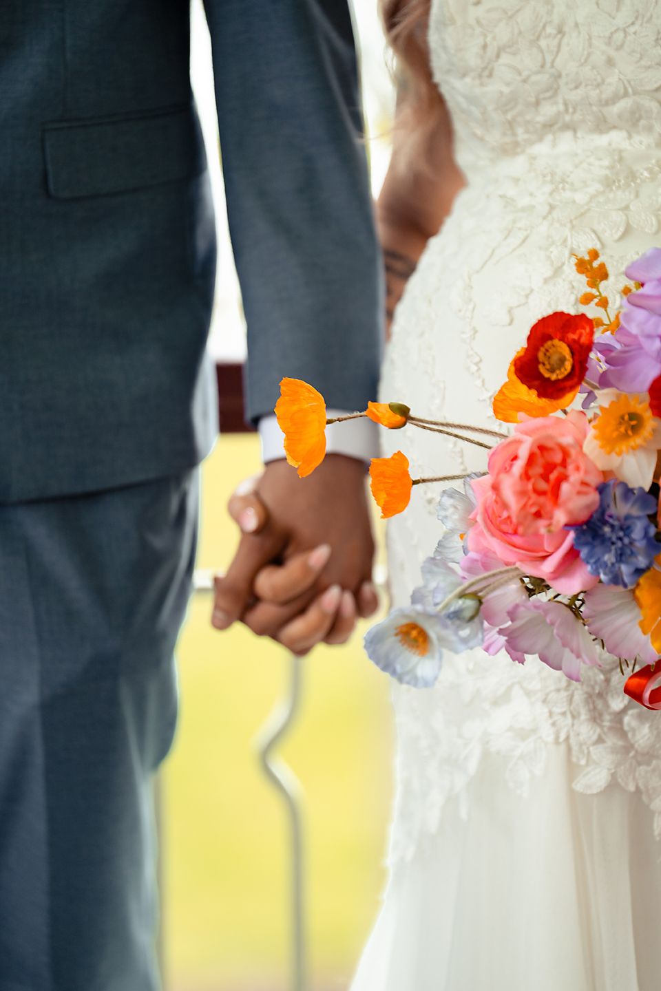 Close-up of newlyweds holding hands with a vibrant bouquet featuring orange and purple flowers.